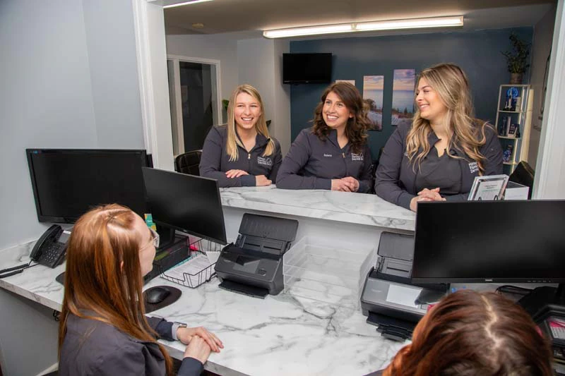 The female staff at Mitten Family Dental standing around the front desk smiling in Ionia MI.